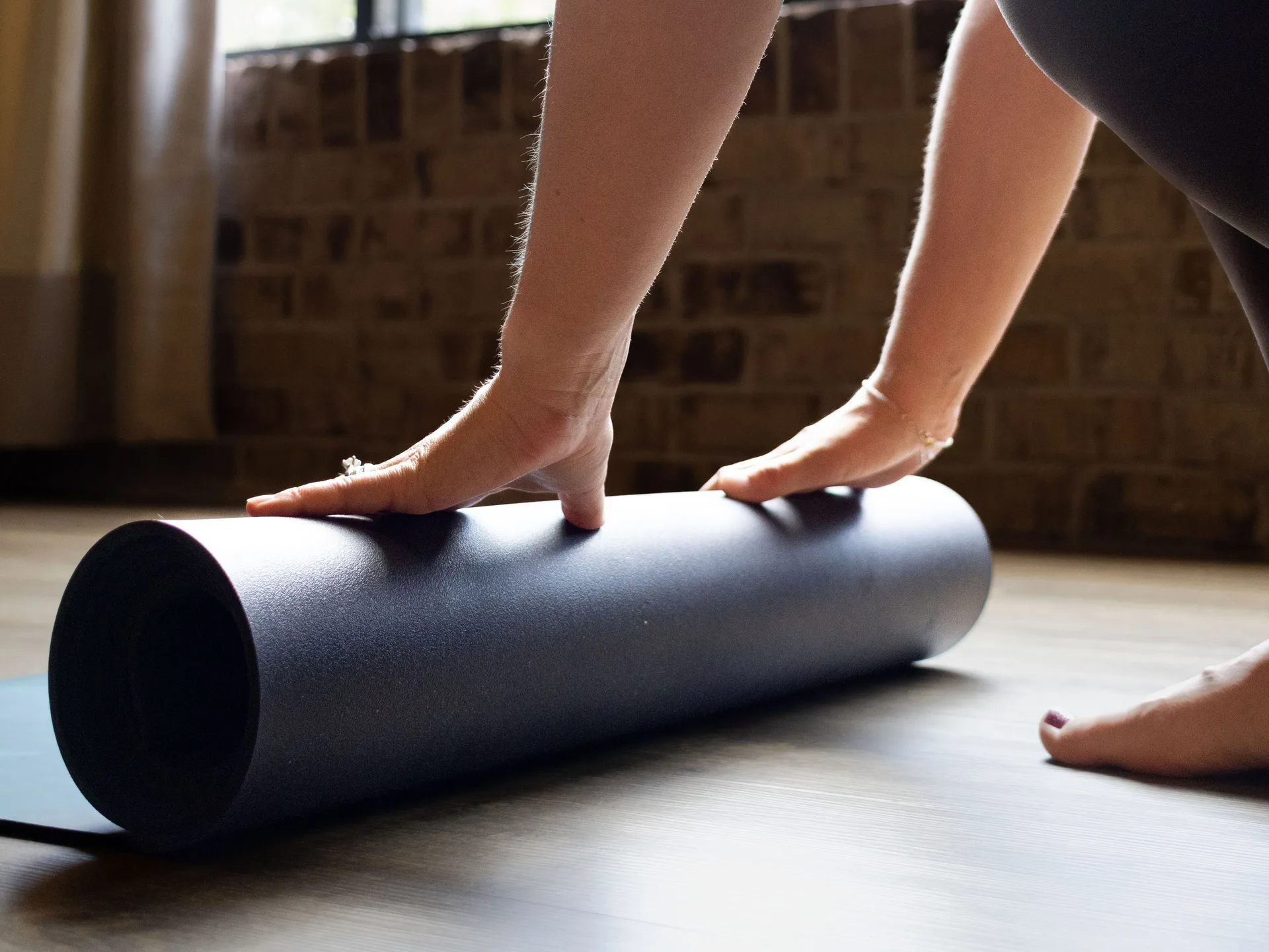 Hands rolling up a yoga mat on the studio floor by the window